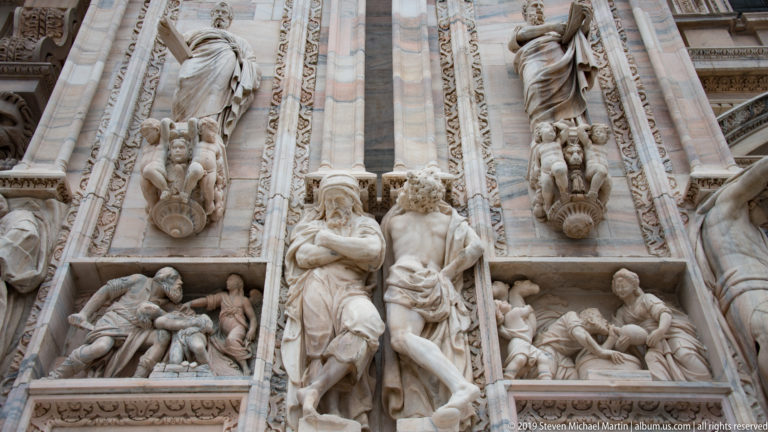 Close-up of carved saints, narrative reliefs, and Gothic architectural ornament on the marble façade of Milan Cathedral.