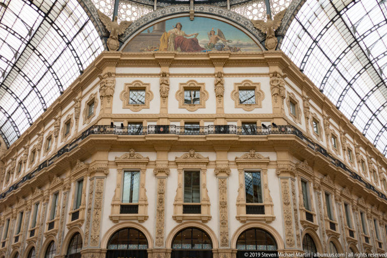 Symmetrical ornate corner façade beneath painted lunettes and the glass roof of the Galleria Vittorio Emanuele II.