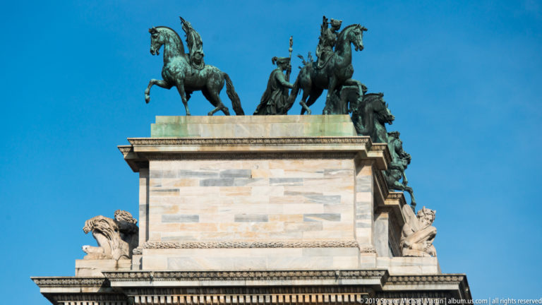Bronze horses and allegorical figures crowning the pale stone Arco della Pace against a blue sky.