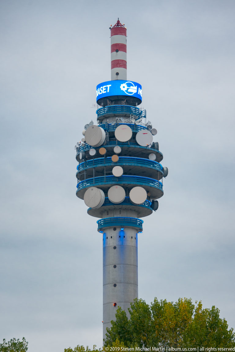 Tall communications tower with stacked circular platforms, many dish antennas, blue bands, and a red-and-white mast.
