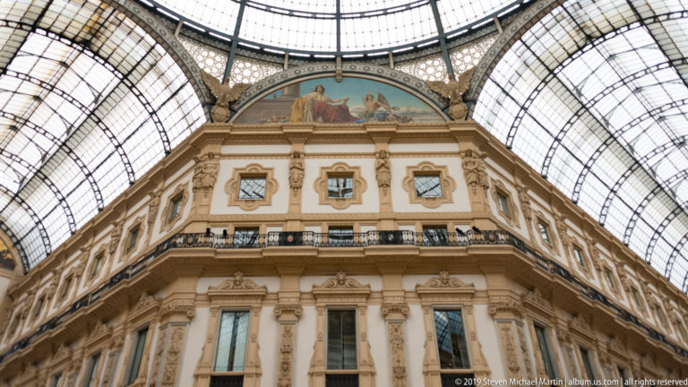 Upward architectural detail of ornate upper façades and glass roof inside the Galleria Vittorio Emanuele II.