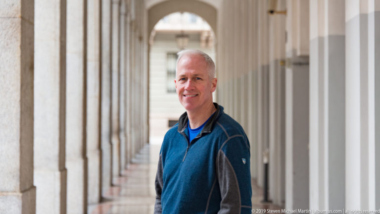 Smiling man in a blue top standing beside a long pale stone colonnade in Milan.