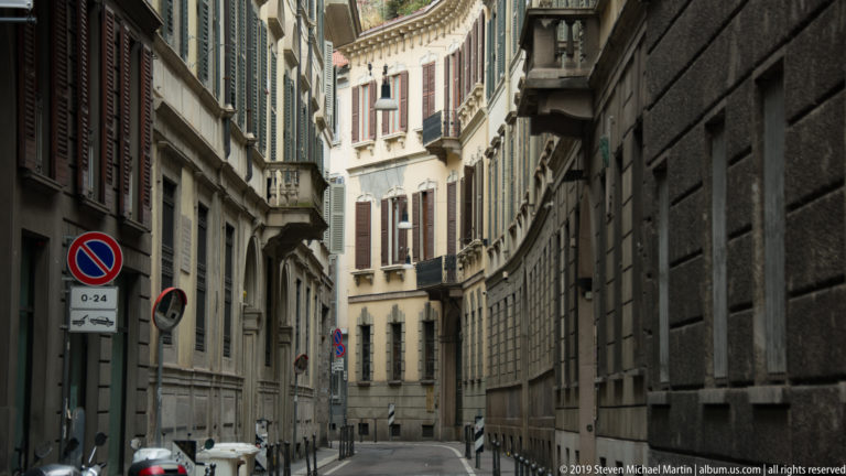 Narrow Milan street lined by tall historic stone buildings, shutters, balconies, parked cars, and traffic signs.