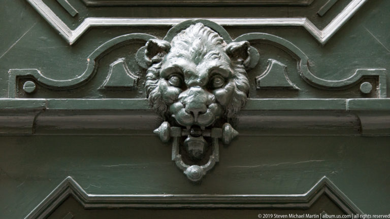 Sculpted lion-head door knocker holding a metal ring on a dark painted panel.