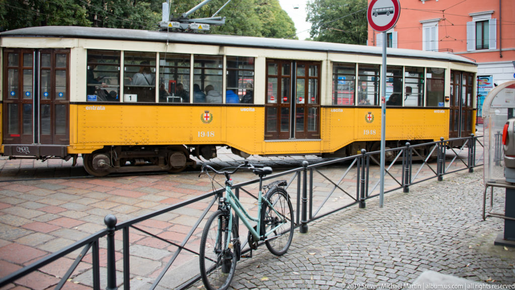 Yellow-orange vintage Milan tram beside a cobbled street, with a bicycle locked to the railing in front.