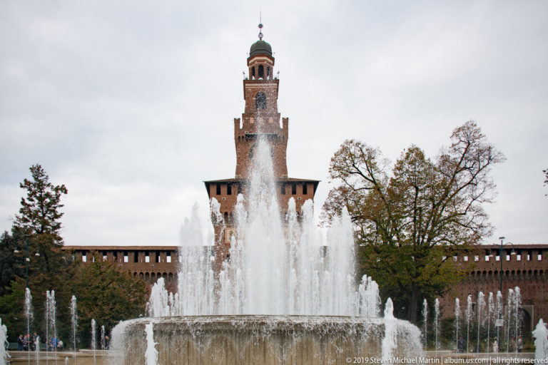 Fountain jets in Piazza Castello with the Torre del Filarete and Sforza Castle walls behind.