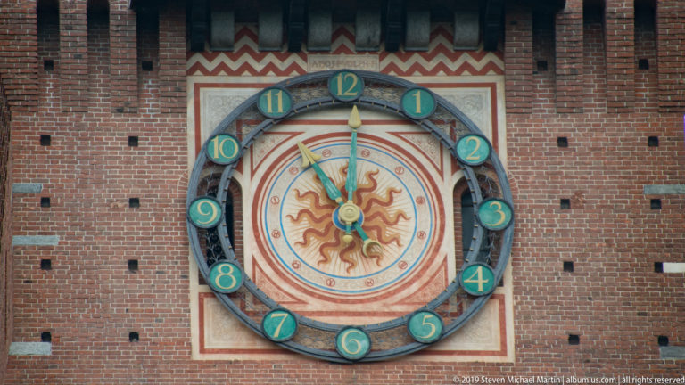 Painted clock face with green numerals and a sun motif on the brick Torre del Filarete at Sforza Castle.