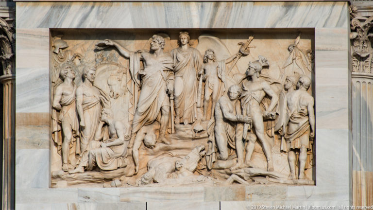 Marble relief on the Arco della Pace showing a crowded classical scene with standing, seated, and kneeling figures.
