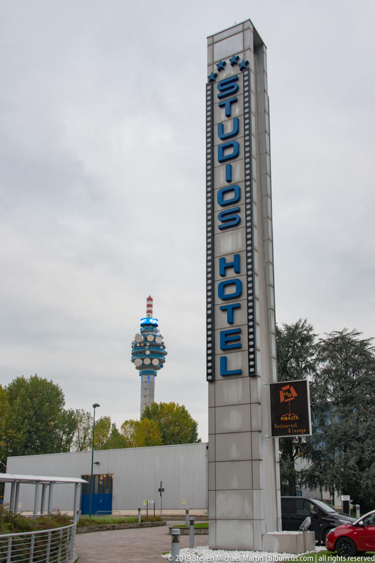 Tall Klima Hotel sign beside a road, with a telecommunications tower rising beyond trees in the background.