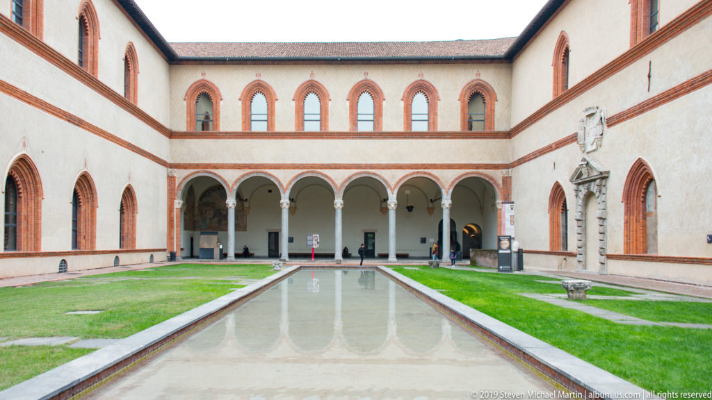 Arcaded courtyard inside Sforza Castle with pale walls, red-brick trim, grass, and a long reflecting pool.