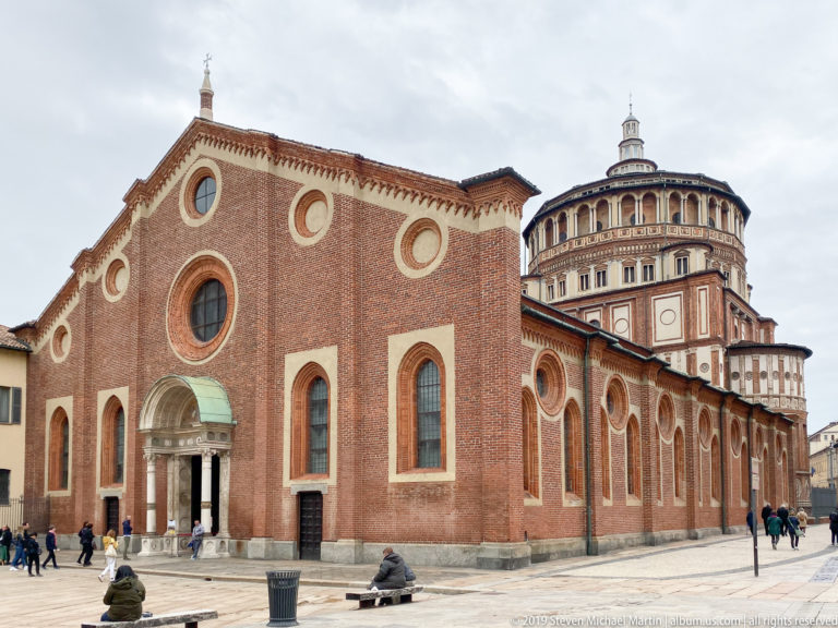Brick exterior of Santa Maria delle Grazie in Milan with round windows and the Renaissance tribune rising behind.