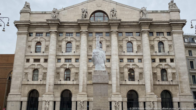 Large pale stone civic building with tall classical columns and a monument centered in the square before it.