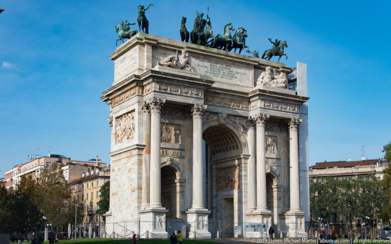 Oblique full view of Milan’s Arco della Pace with three arches, Corinthian columns, relief panels, and bronze sculpture above.