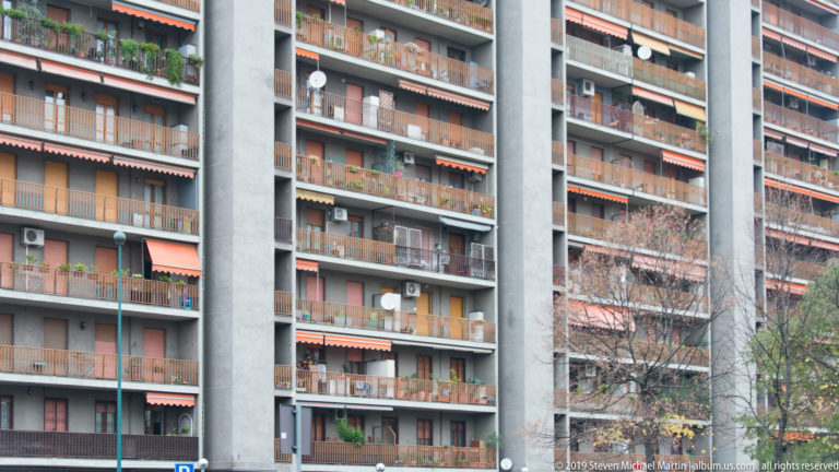 Modern Milan apartment building with stacked balconies, gray concrete piers, awnings, plants, and bare tree branches.