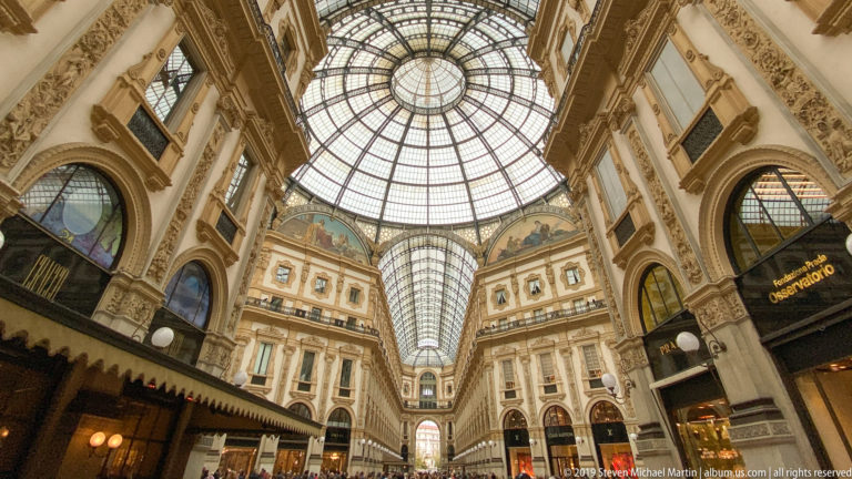 Axial interior view through the Galleria Vittorio Emanuele II with glass vaults, dome, shops, and pedestrians.