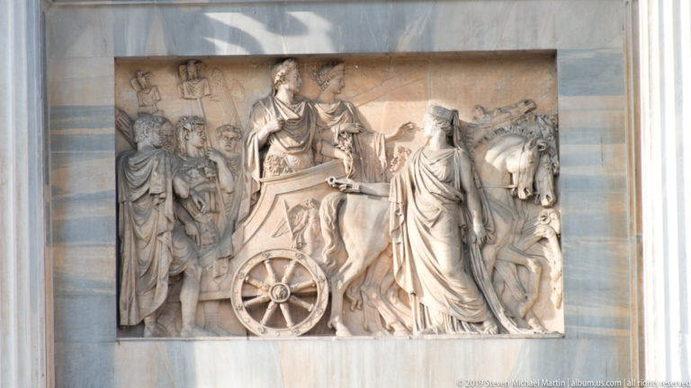 Marble relief on the Arco della Pace with a chariot, draped figures, attendants, and a horse at right.