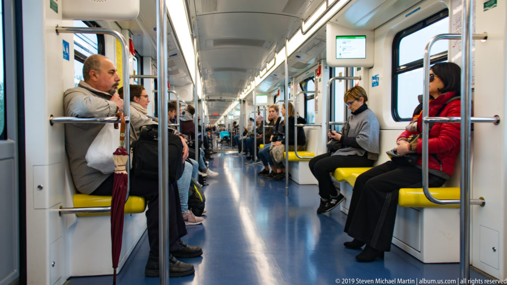 Passengers seated on yellow benches inside a bright Milan Metro carriage with metal grab poles along the aisle.