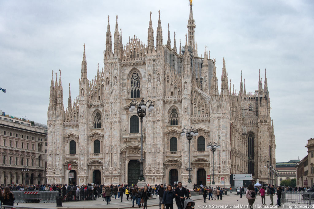 Wide view of Milan Cathedral’s marble façade and spired roofline with pedestrians crossing Piazza del Duomo in the foreground.