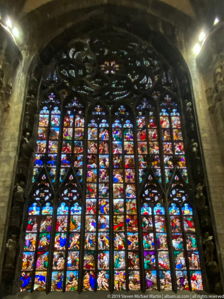 Large arched stained-glass window glowing inside the cathedral, with dense narrative panels below elaborate dark Gothic tracery.