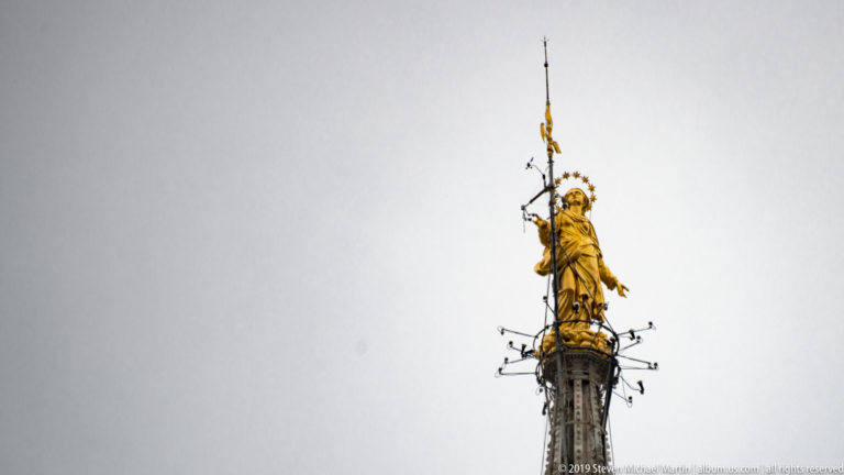 Distant gilded Madonnina with outstretched arm and halo atop a slender spire against a broad gray sky.
