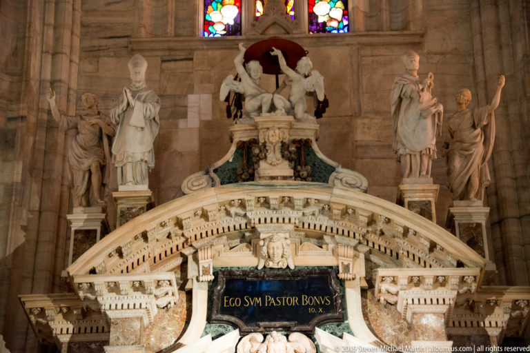 Ornate marble monument with standing figures, a miniature architectural panorama, and the inscription “EGO SUM PASTOR BONUS.”