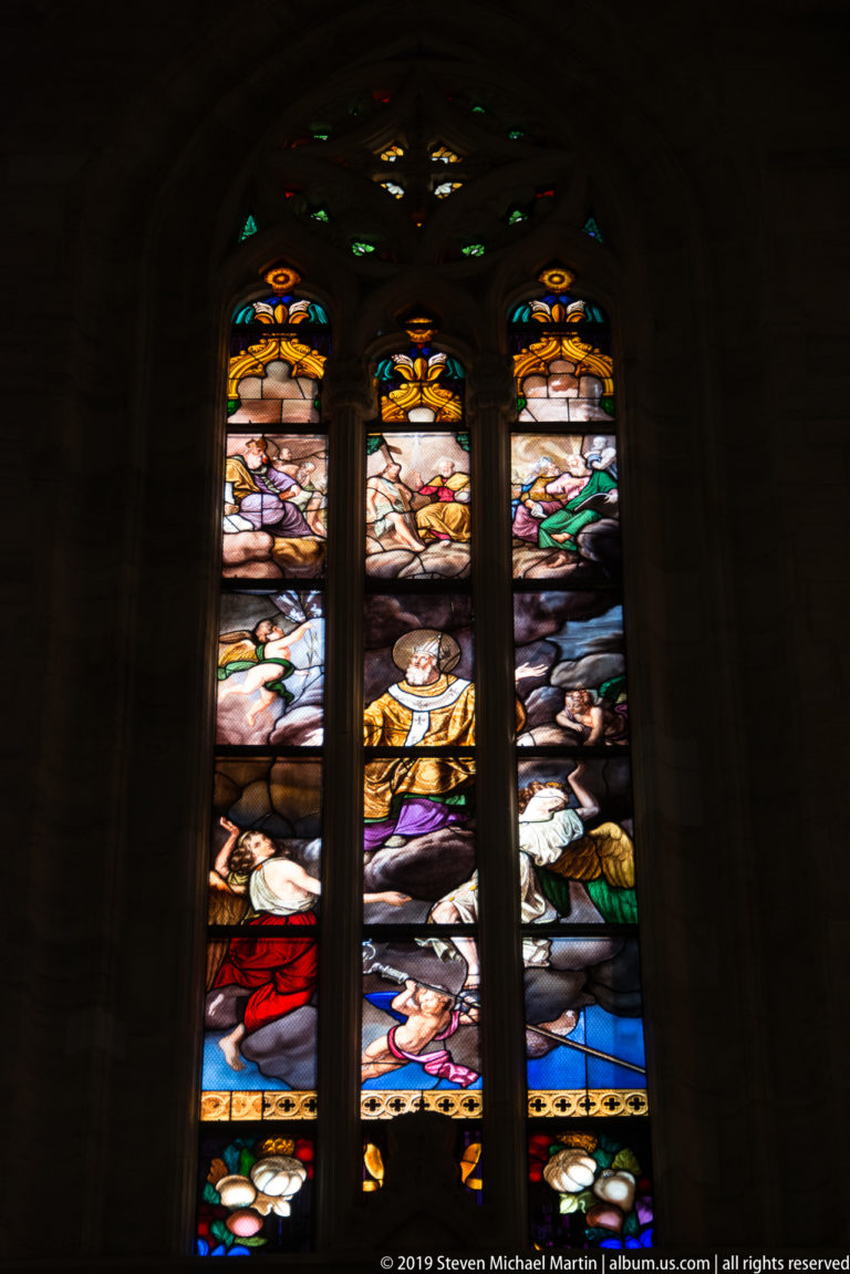 Three narrow stained-glass lancets glow in a dark interior, each filled with stacked colorful biblical scenes.
