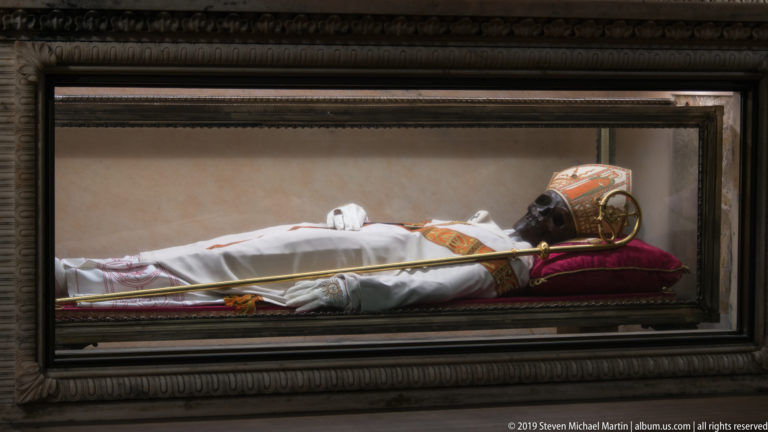 Venerated remains of Saint Charles Borromeo displayed in a glass reliquary, dressed in episcopal vestments and mitre.