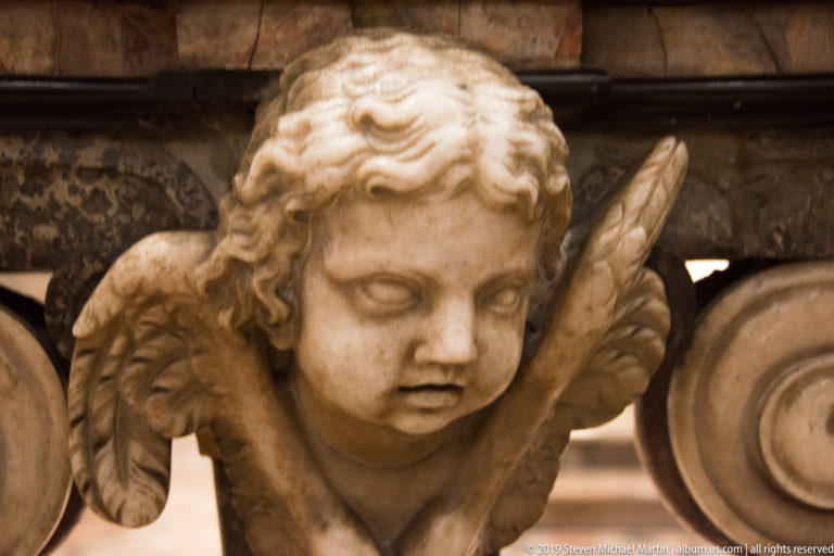 Close view of a carved marble cherub head with curled hair and feathered wings on an interior monument.