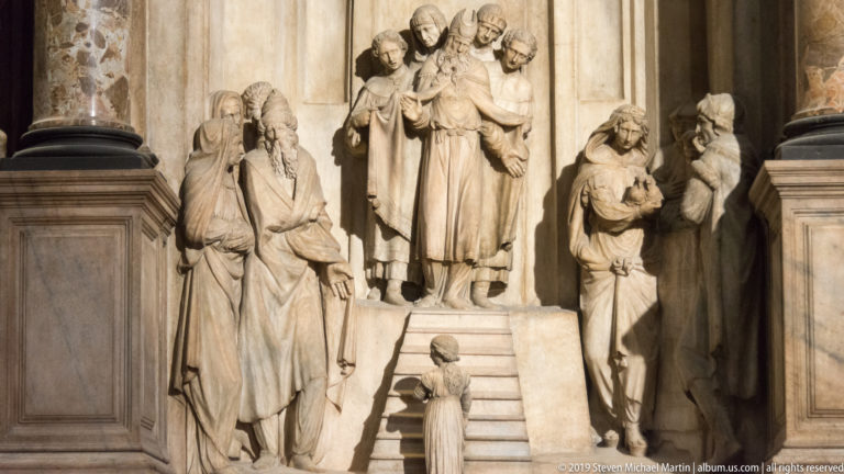 Marble monument with robed clerics and attendants grouped around a short stair, including a small figure at the base.