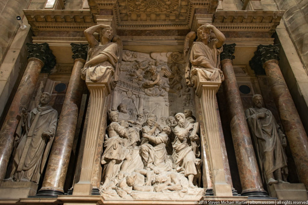 Tall marble wall monument with a crowded central relief, colored columns, seated figures above, and standing figures at the sides.