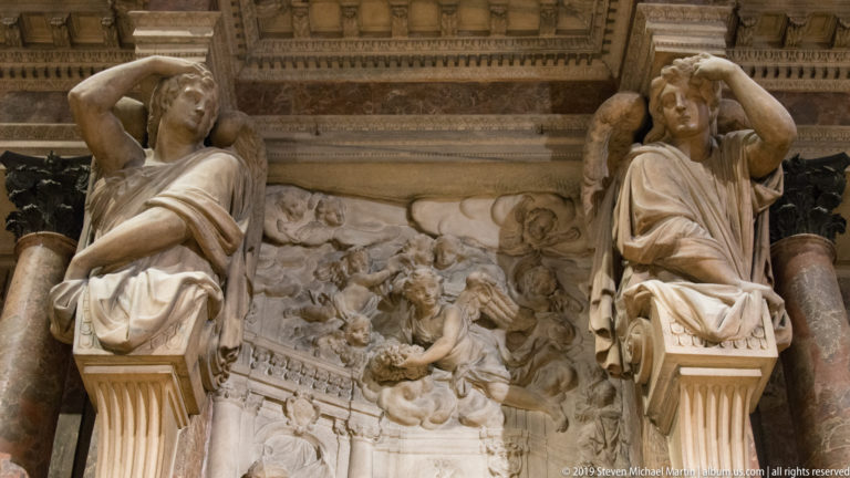 Two seated marble figures flank a crowded relief above colored columns and ornate architectural moldings.