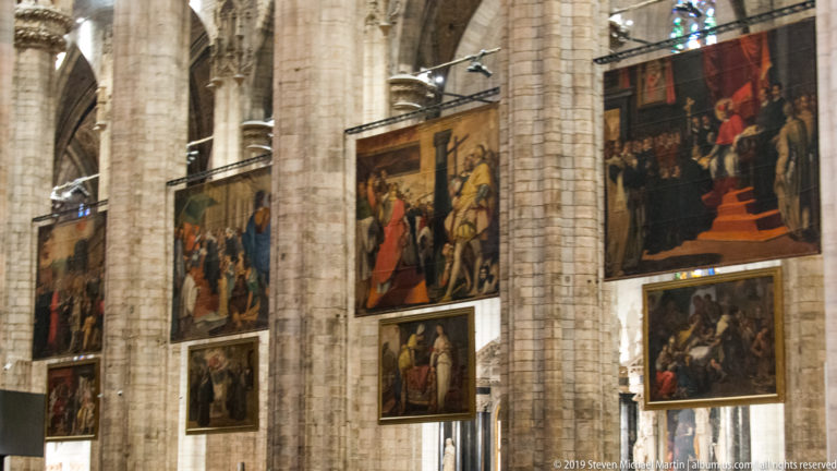 Large Quadroni di San Carlo canvases hanging between massive Gothic piers inside the Duomo di Milano.