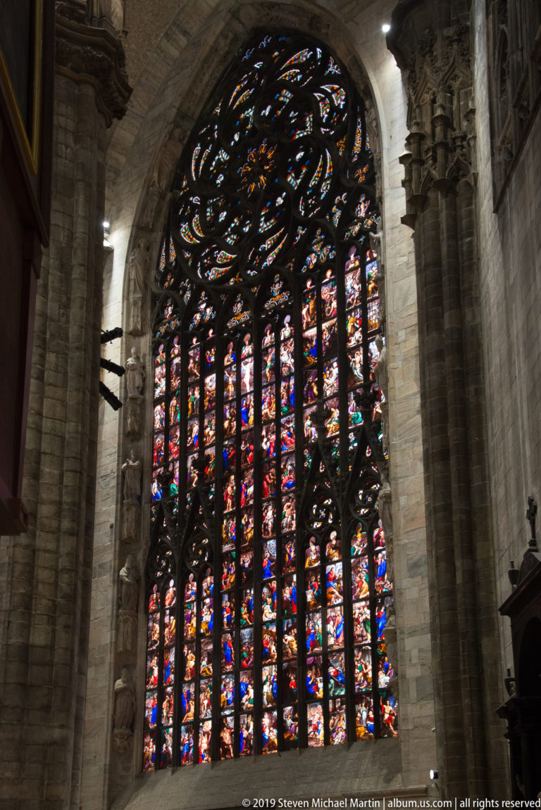 Large stained-glass window glows within a deep stone arch, framed by massive cathedral piers and dark Gothic tracery.