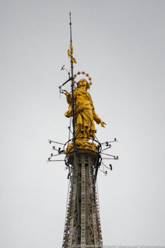 Gilded Madonnina statue with starry halo atop Milan Cathedral’s highest spire, surrounded by protective metalwork.