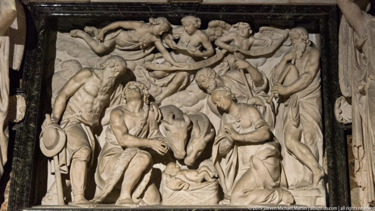 Marble Nativity relief with Mary and Joseph beside the infant Jesus, flanked by visitors and angels overhead.