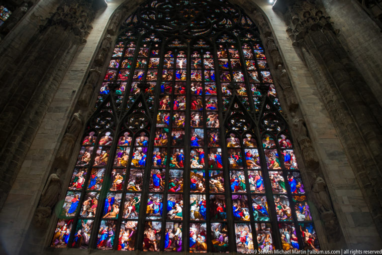 Tall stained-glass window rises inside a pointed stone arch, with colorful narrative panels below intricate dark tracery.