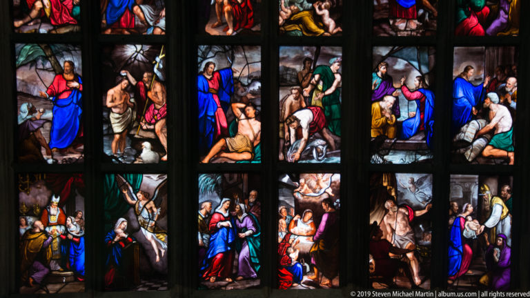 Stained-glass detail with alternating standing figures and crowded biblical scenes separated into narrow vertical compartments.