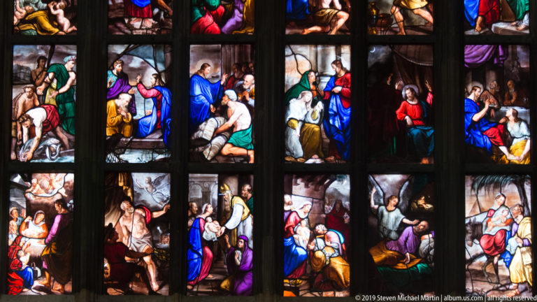 Close stained-glass panels contain compact biblical scenes with figures gathered in interiors and around beds or doorways.