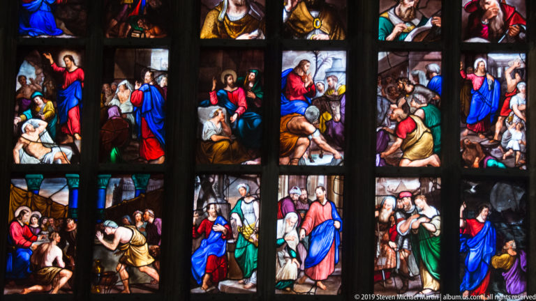 Colorful stained-glass scenes show standing, kneeling, and seated biblical figures in narrow panels divided by dark mullions.