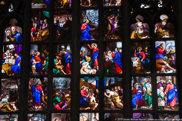 Vertical stained-glass bays contain colorful biblical scenes, with small arched figure panels crowning the narrative sequence.