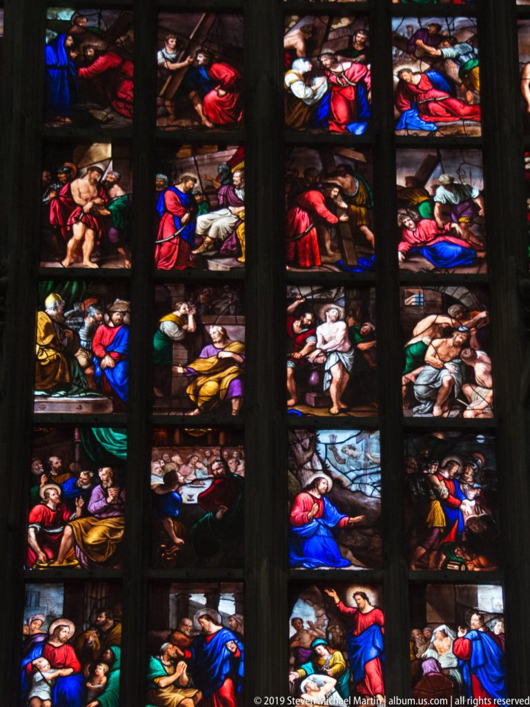 Close stained-glass view with stacked biblical scenes and vividly robed figures separated by heavy dark vertical bars.