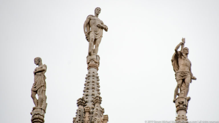 Three stone statues standing atop ornate Gothic pinnacles against a pale overcast sky.
