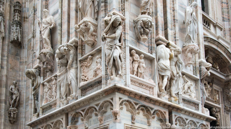 Oblique cathedral corner covered with standing statues, crouching supports, narrative reliefs, carved borders, and pointed arcading.
