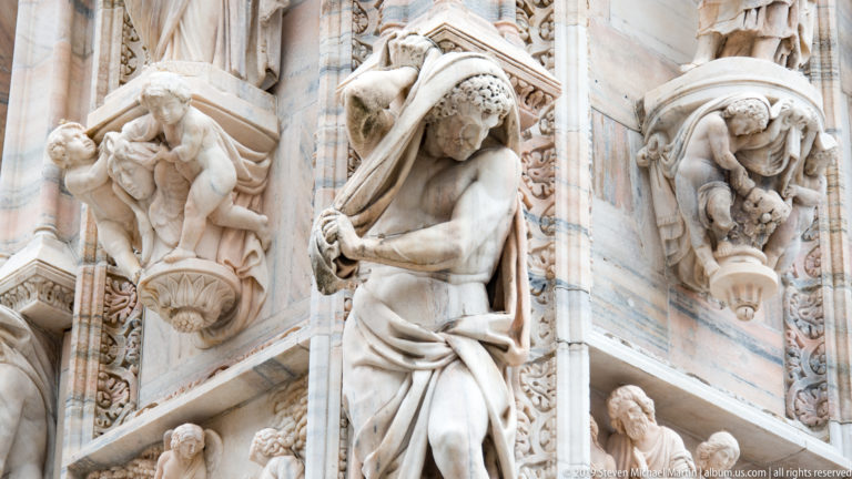 Close view of pale marble exterior carving with a draped standing figure, crouching figures, grotesques, foliage, and projecting supports.