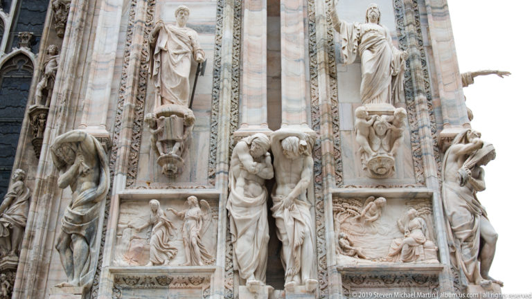 Vertical façade bay combining standing statues, crouching supporting figures, narrative relief panels, and dense carved borders.