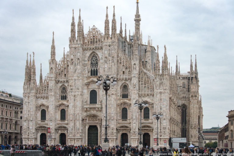 Frontal view of Milan Cathedral’s ornate marble west façade and spires with crowds gathered across Piazza del Duomo.