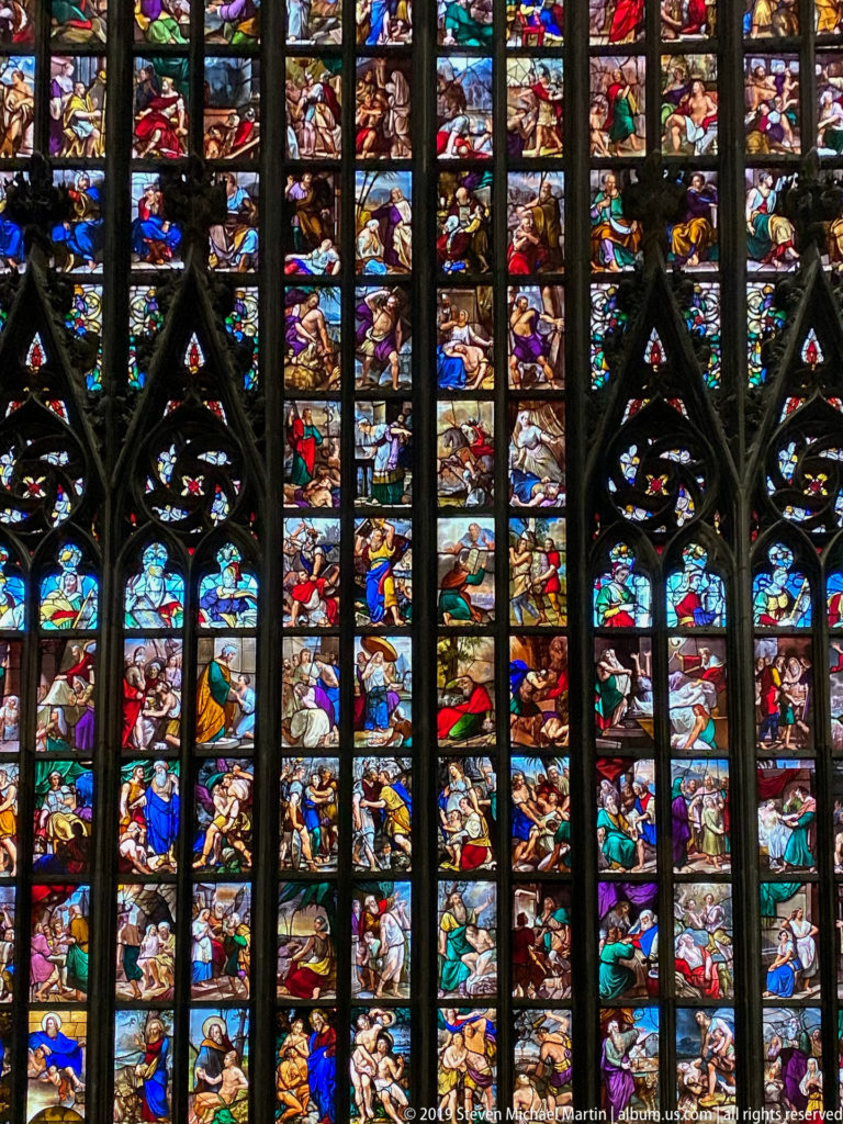 Colorful biblical figures and scenes fill stacked stained-glass panels divided by dark Gothic mullions and tracery.