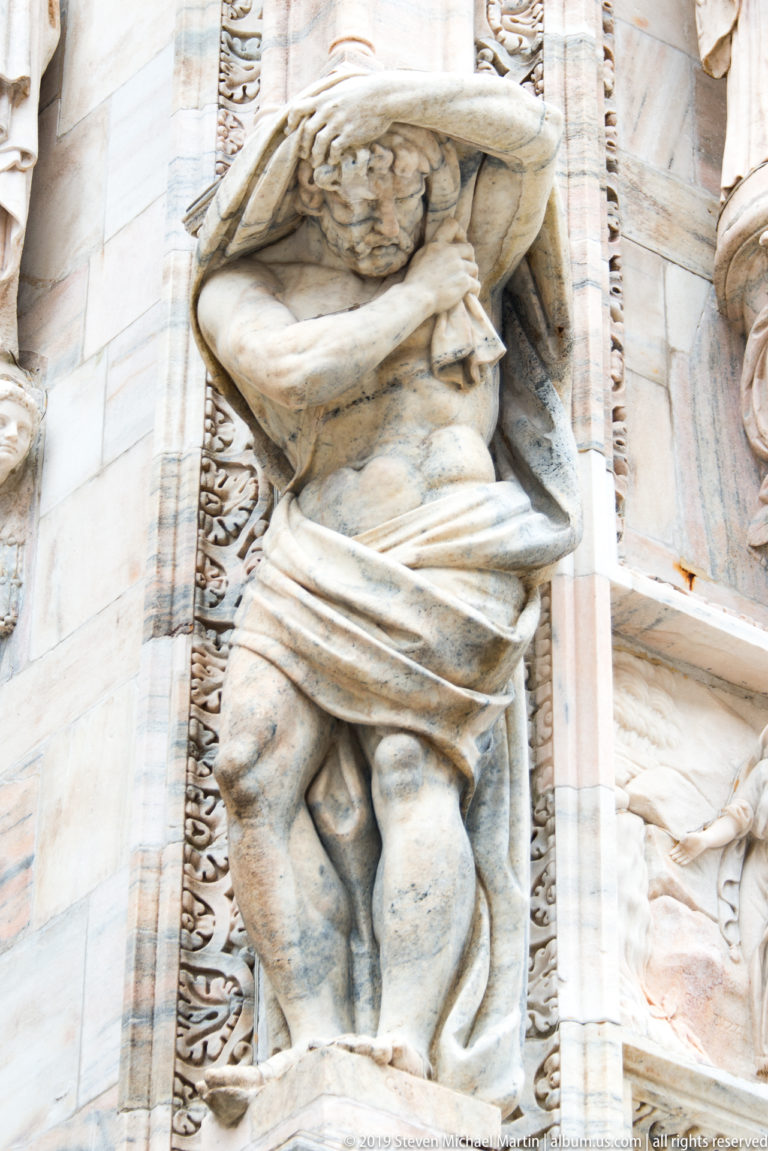 Full crouching marble figure with raised arms supporting a projecting cornice, framed by carved borders and marble masonry.