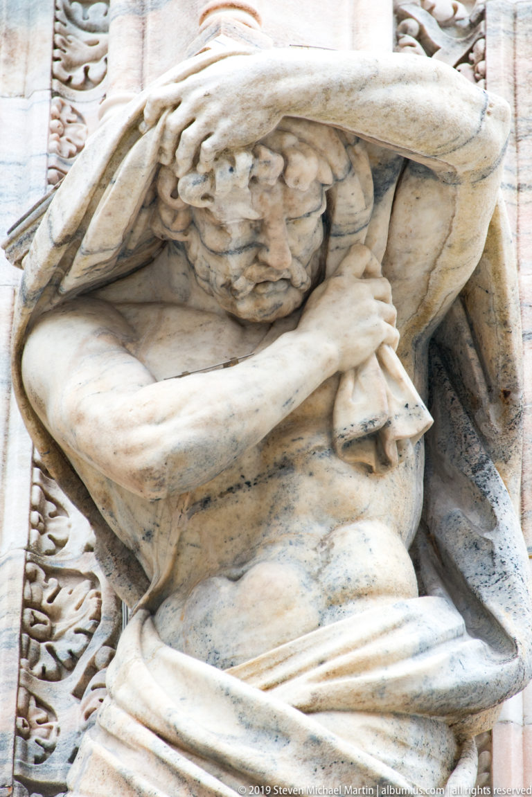 Crouching muscular marble figure bracing beneath a heavy architectural projection on the cathedral façade.