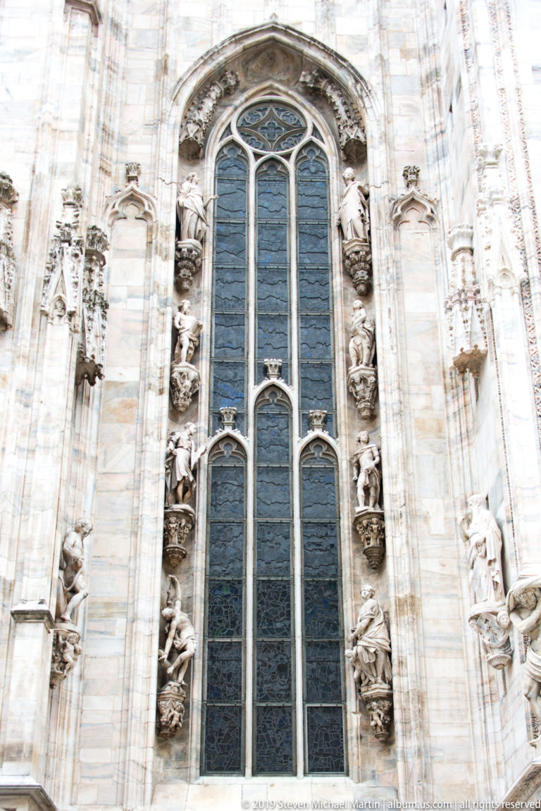 Tall pointed Gothic window with dark glazing, vertical mullions, curving tracery, and rows of statues along both sides.